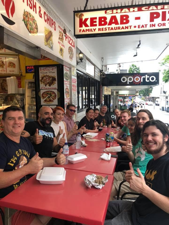 A group of customers giving thumbs up over takeaway boxes at the tables outside SHAH Kebabs