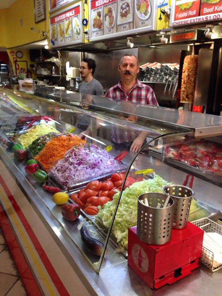 The SHAH Kebabs counter filled with shredded lettuce, tomato, onion, carrot and parsley salad, with our team standing behind it